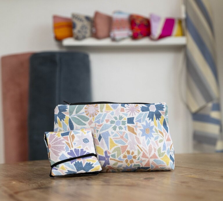 Zwei Kosmetiktaschen mit floralen Mustern in Pastellfarben stehen auf einem Holztisch, im Hintergrund Stoffe und Deko. | © BILD der FRAU