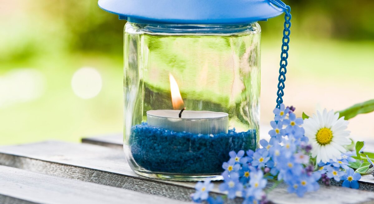 Ein Windlicht mit blauer Metallhaube und brennender Kerze steht auf einem Holztisch, umgeben von blauen Blüten.