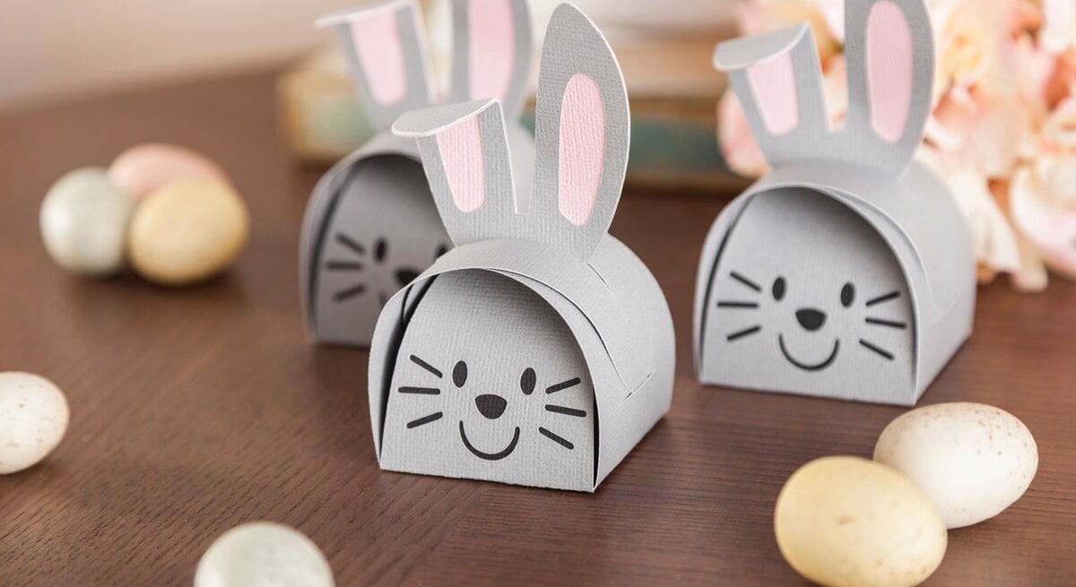 Drei graue Osterhasen-Geschenkboxen mit rosa Ohren und aufgemalten Gesichtern stehen auf einem Holztisch, umgeben von bunten Eiern.