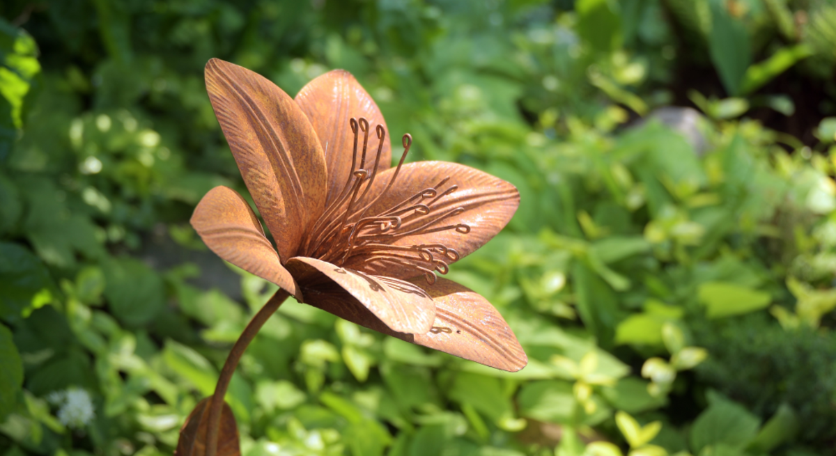 Rostfarbene Metallblume mit filigranen Staubblättern steht kunstvoll gebogen inmitten dichter grüner Gartenpflanzen.