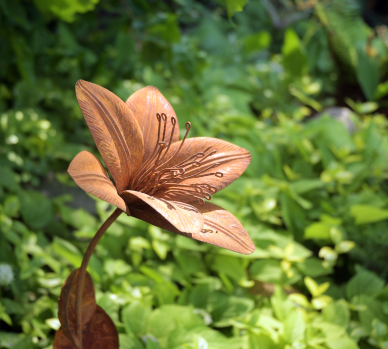 Rostfarbene Metallblume mit filigranen Staubblättern steht kunstvoll gebogen inmitten dichter grüner Gartenpflanzen. | © AdobeStock/Kalle Kolodziej
