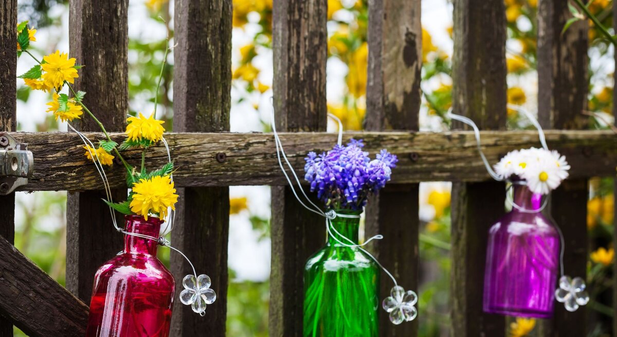 Drei bunte Glasflaschen mit gelben, blauen und weißen Blumen hängen dekorativ an einem alten Holzzaun im blühenden Garten.