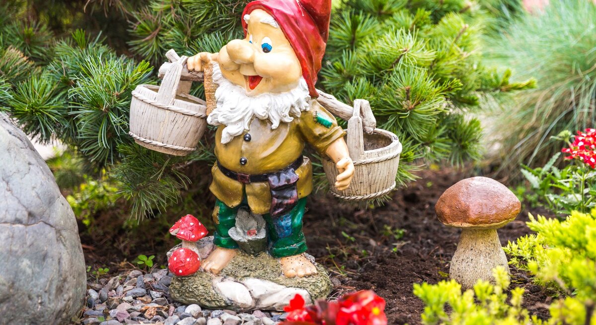 Ein bunter Gartenzwerg mit roter Mütze und weißen Bart trägt zwei Holzeimer und steht lachend zwischen grünen Pflanzen und Pilzen.
