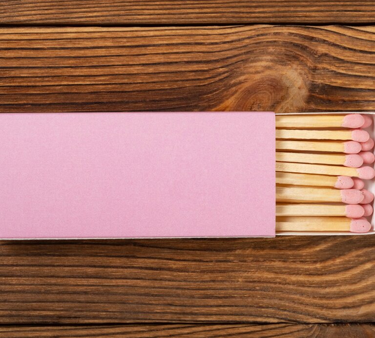 Eine halb geöffnete Streichholzschachtel mit rosa Zündköpfen liegt auf einer rustikalen Holzoberfläche. | © Adobe Stock/Freer