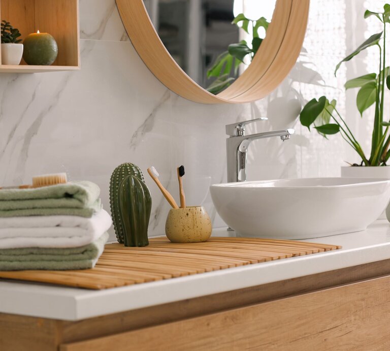 Modernes Badezimmer mit weißem Waschbecken, Holzdetails, grünen Pflanzen und gefalteten Handtüchern auf einer Bambusmatte. | © Adobe Stock/New Africa