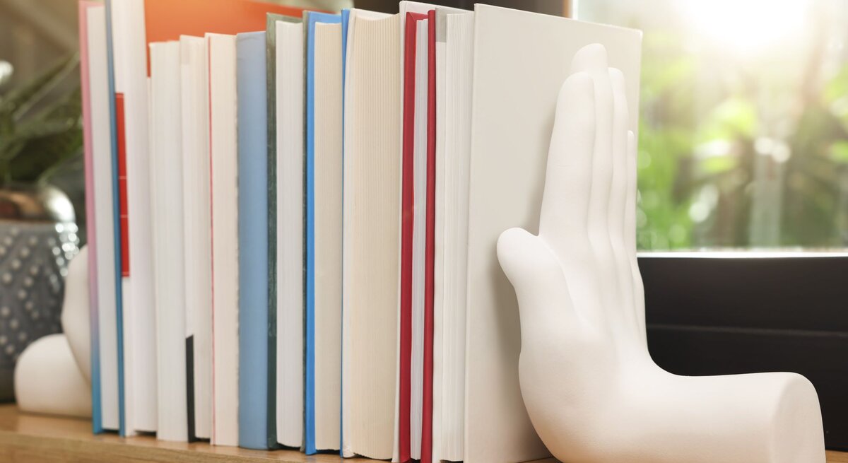 Eine Reihe bunter Bücher steht auf einem Holzregal, gestützt von weißen Buchstützen in Form einer Hand vor einem sonnigen Fenster.
