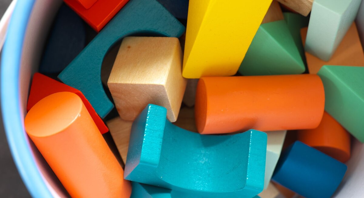 Ein Eimer voller bunter Holzbausteine in verschiedenen Formen, darunter Zylinder, Würfel und Dreiecke in Blau, Orange, Gelb und Grün.