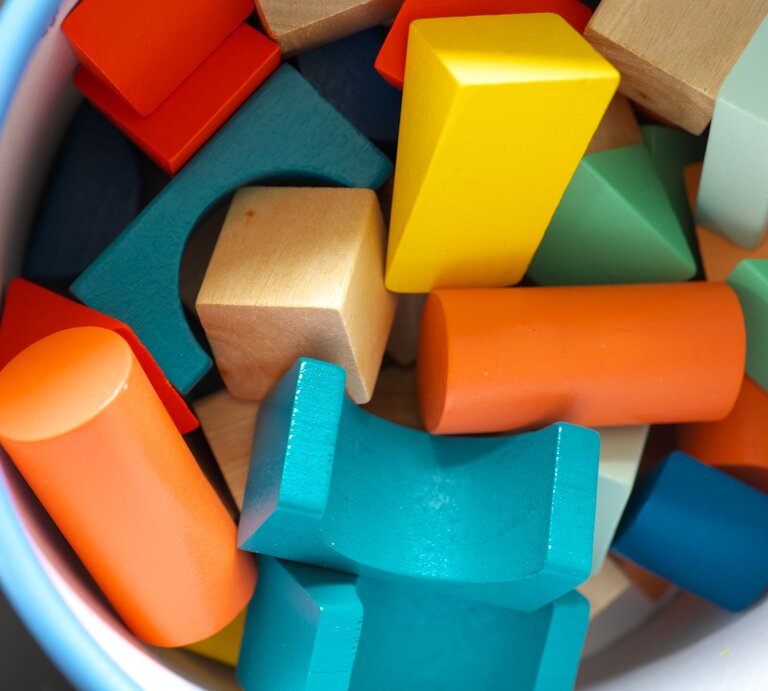 Ein Eimer voller bunter Holzbausteine in verschiedenen Formen, darunter Zylinder, Würfel und Dreiecke in Blau, Orange, Gelb und Grün. | © Adobe Stock/Udo Bojahr