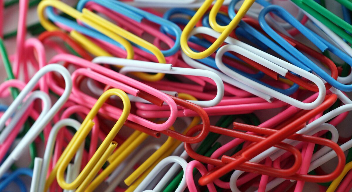 Ein chaotischer Haufen bunter Büroklammern in Gelb, Blau, Rot, Grün, Weiß und Pink, teilweise ineinander verhakt.