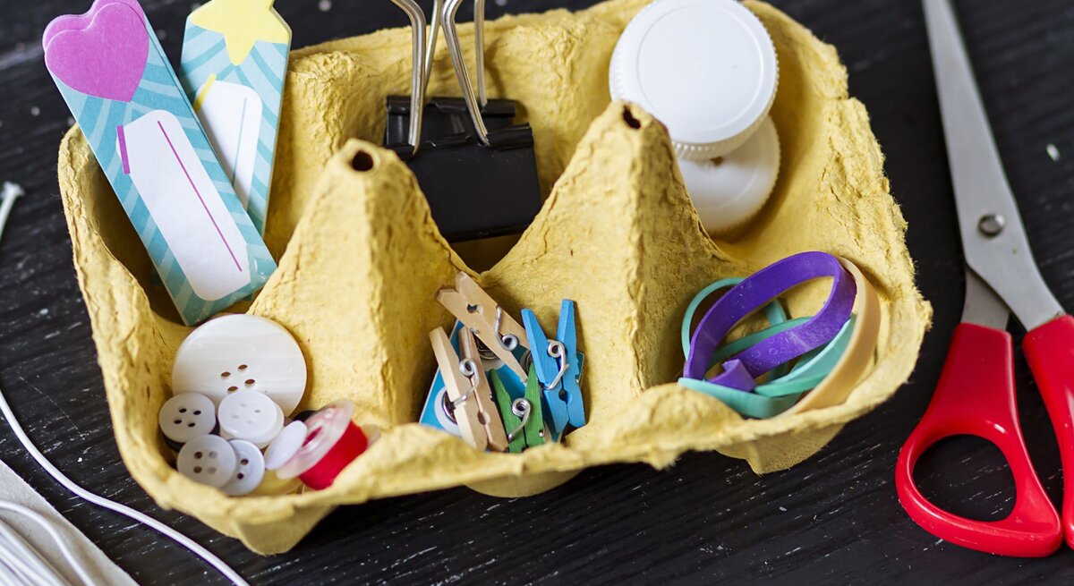 Eine gelbe Eierkartonbox dient als Aufbewahrungsbox für Bastelmaterialien wie Knöpfe, Gummibänder, Büroklammern und bunte Holzklammern.
