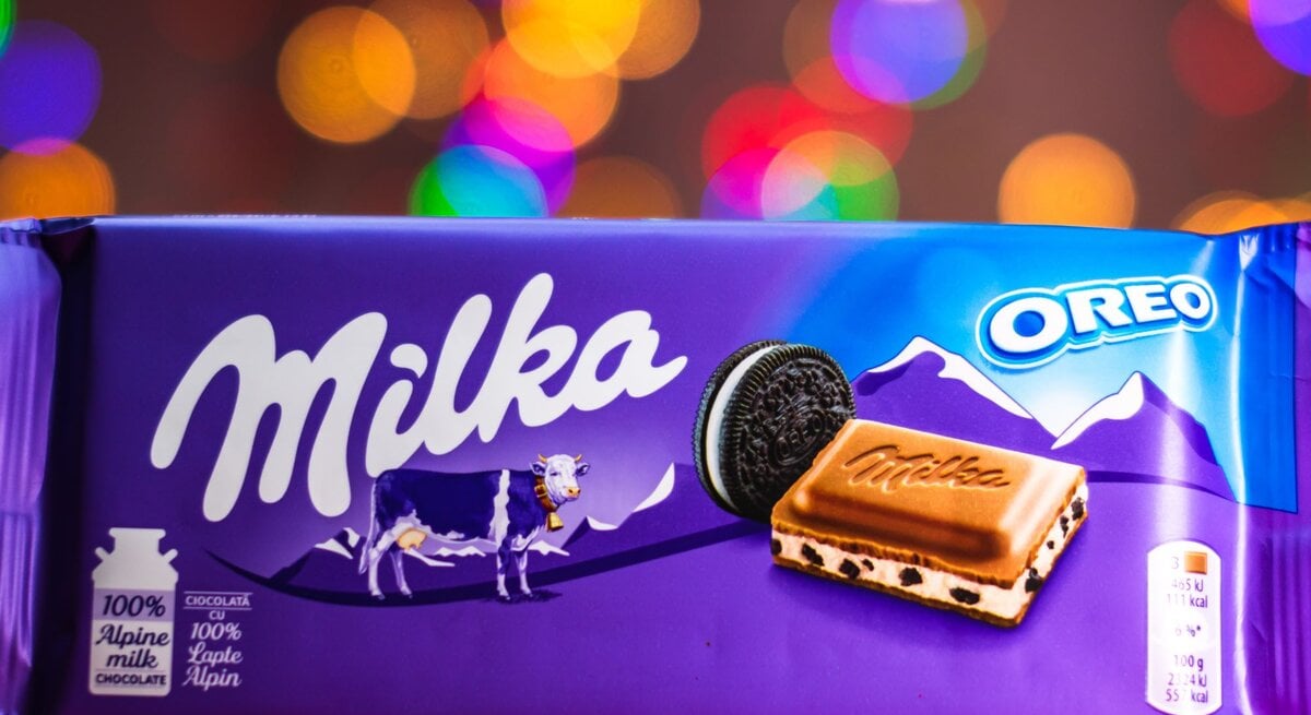 Eine Tafel Milka-Oreo-Schokolade in lilafarbener Verpackung mit Alpenmotiv und Milka-Kuh, vor buntem, unscharfem Lichterhintergrund.