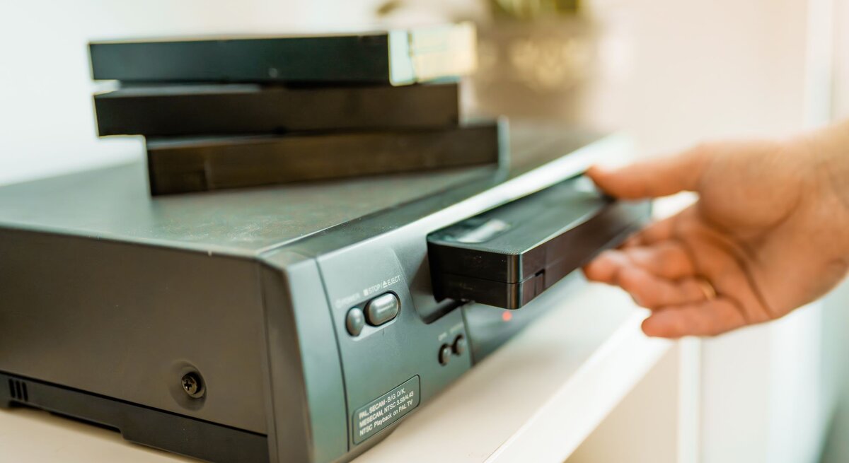 Nahaufnahme eines VHS-Players, in den eine Hand eine Kassette einlegt, während mehrere schwarze Videokassetten gestapelt darauf liegen.