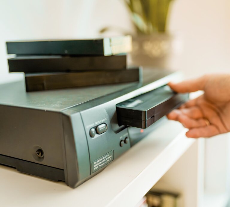 Nahaufnahme eines VHS-Players, in den eine Hand eine Kassette einlegt, während mehrere schwarze Videokassetten gestapelt darauf liegen. | © Adobe Stock/Victoria