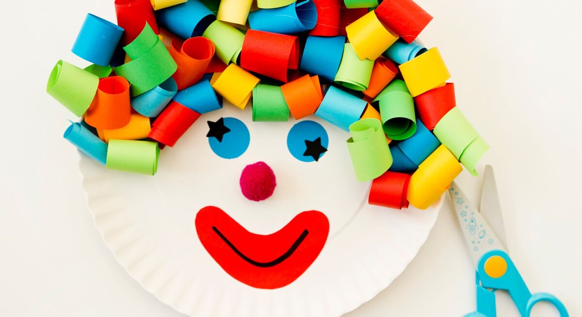Buntes Clown-Gesicht aus einem Pappteller mit Locken aus gerolltem Papier, roten Lippen und Pompon-Nase, daneben eine Schere und eine Kinderhand.