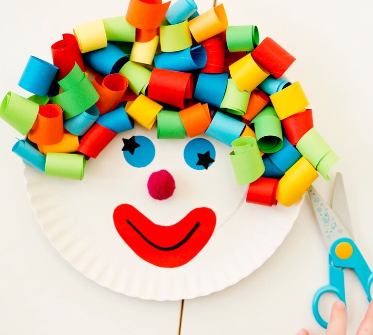 Buntes Clown-Gesicht aus einem Pappteller mit Locken aus gerolltem Papier, roten Lippen und Pompon-Nase, daneben eine Schere und eine Kinderhand. | © Adobe Stock/Birute