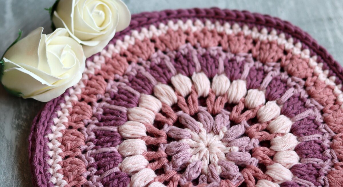 Ein Topfuntersetzer als handgefertigte Häkelarbeit mit kreisförmigem Farbverlauf in Rosa und Lila, ergänzt durch zwei edle weiße Rosen auf Betonhintergrund.