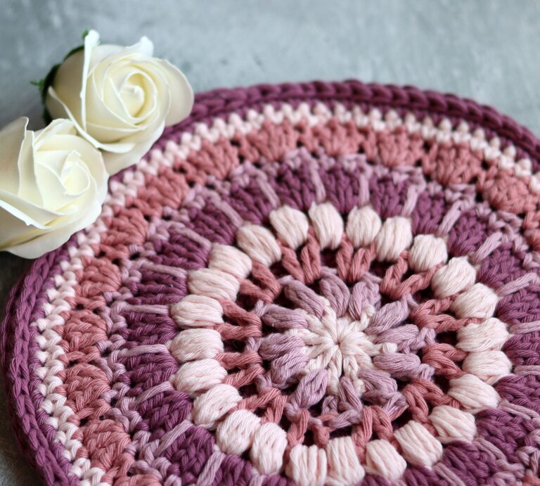 Ein Topfuntersetzer als handgefertigte Häkelarbeit mit kreisförmigem Farbverlauf in Rosa und Lila, ergänzt durch zwei edle weiße Rosen auf Betonhintergrund. | © Adobe Stock/Maya