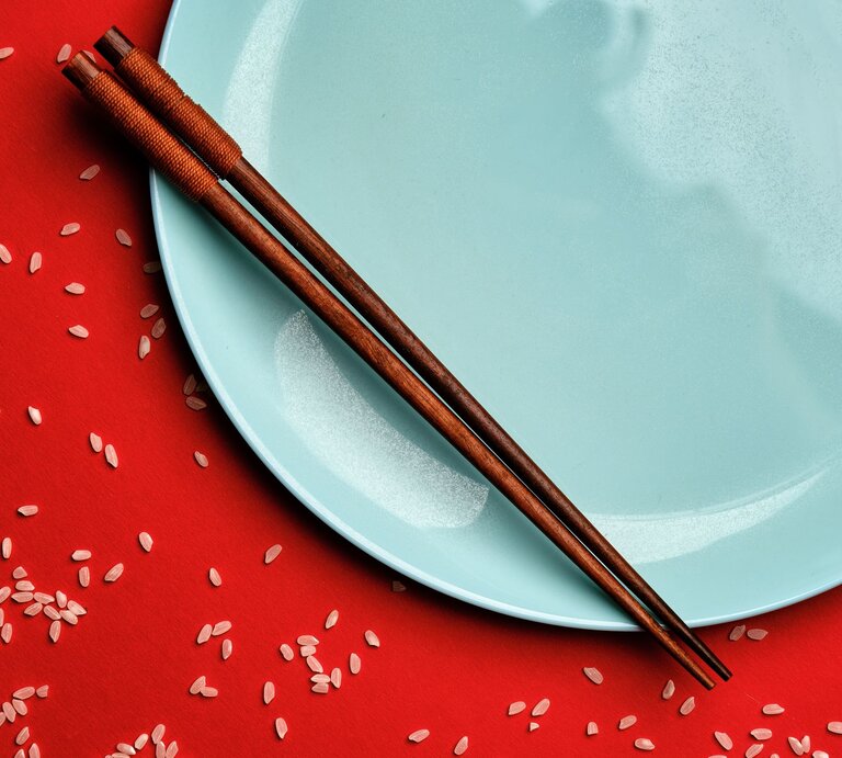 Zwei elegante Essstäbchen aus Holz auf einem glänzenden Teller, daneben lose Reiskörner auf kräftigem Rot als Kontrast. | © Adobe Stock/vladimirkolens