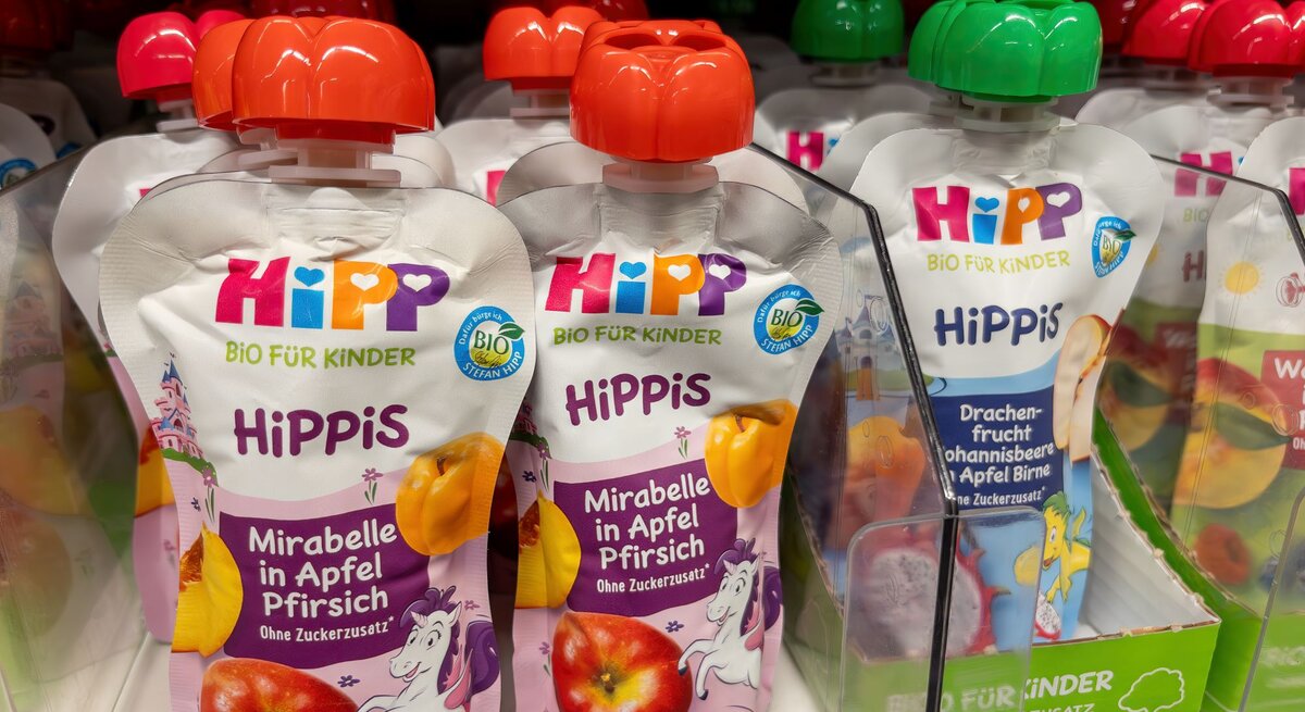 Mehrere HiPP-Quetschbeutel mit Fruchtpüree für Kinder stehen im Regal, vorn die Sorte Mirabelle in Apfel Pfirsich mit lila Einhorn-Design.
