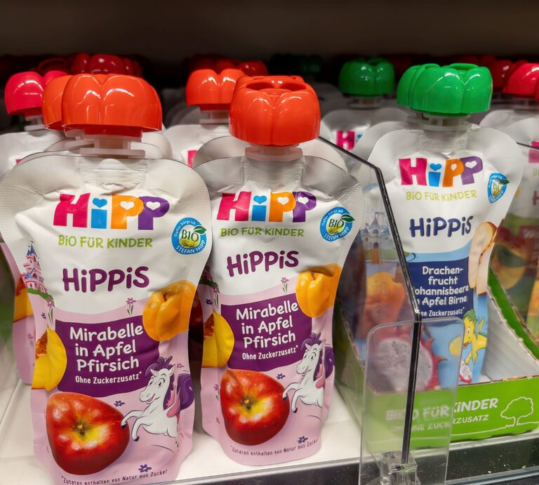 Mehrere HiPP-Quetschbeutel mit Fruchtpüree für Kinder stehen im Regal, vorn die Sorte Mirabelle in Apfel Pfirsich mit lila Einhorn-Design. | © Adobe Stock/VGV