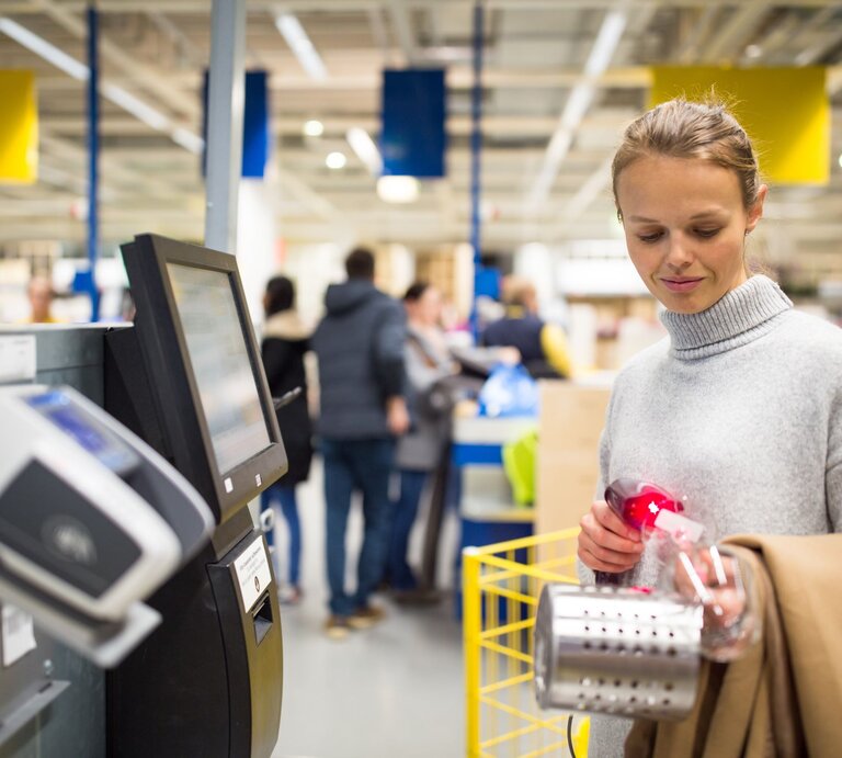 Eine junge Frau mit hellbraunen Haaren im grauen Rollkragenpullover scannt an einer Ikea-SB-Kasse einen Besteckständer, im Hintergrund ein geschäftiger Laden. | © Adobe Stock/lightpoet