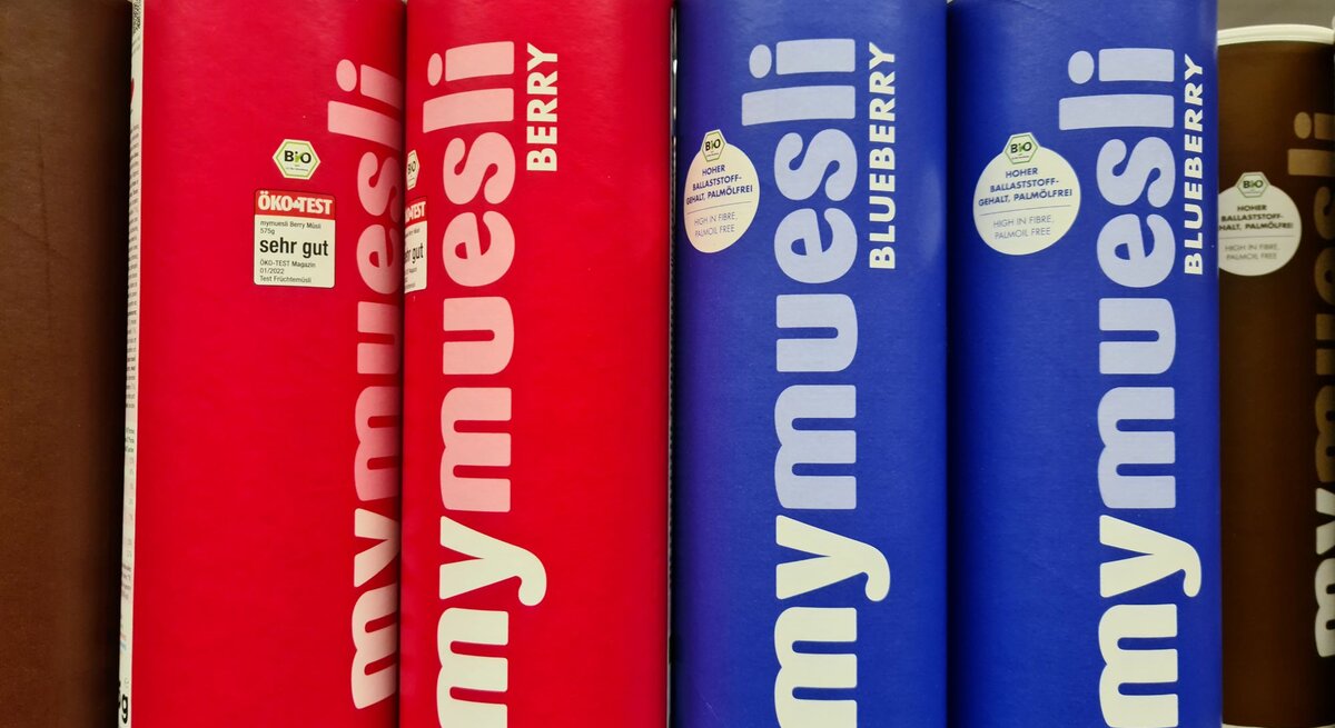 Vier Müslidosen der Marke mymuesli, zwei in Rot für Beerenmüsli und zwei in Blau für Blaubeermüsli, stehen nebeneinander im Regal.