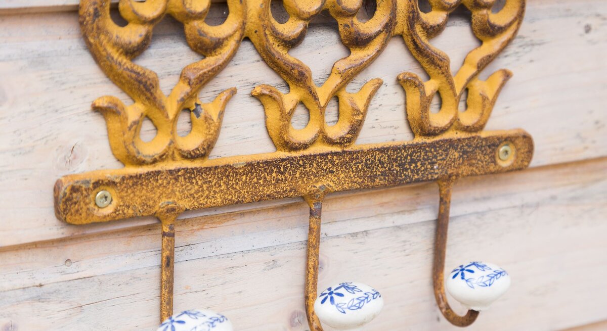 Drei antike, goldgelbe Haken mit filigranen Verzierungen und weißen Keramikknäufen mit blauen Blumenmotiven an einer Holzlattenwand.