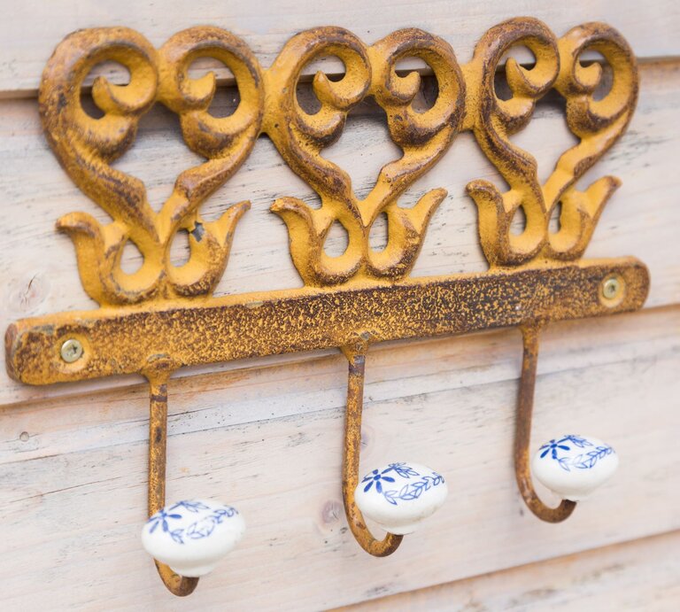 Drei antike, goldgelbe Haken mit filigranen Verzierungen und weißen Keramikknäufen mit blauen Blumenmotiven an einer Holzlattenwand. | © Adobe Stock/Bernd Kröger