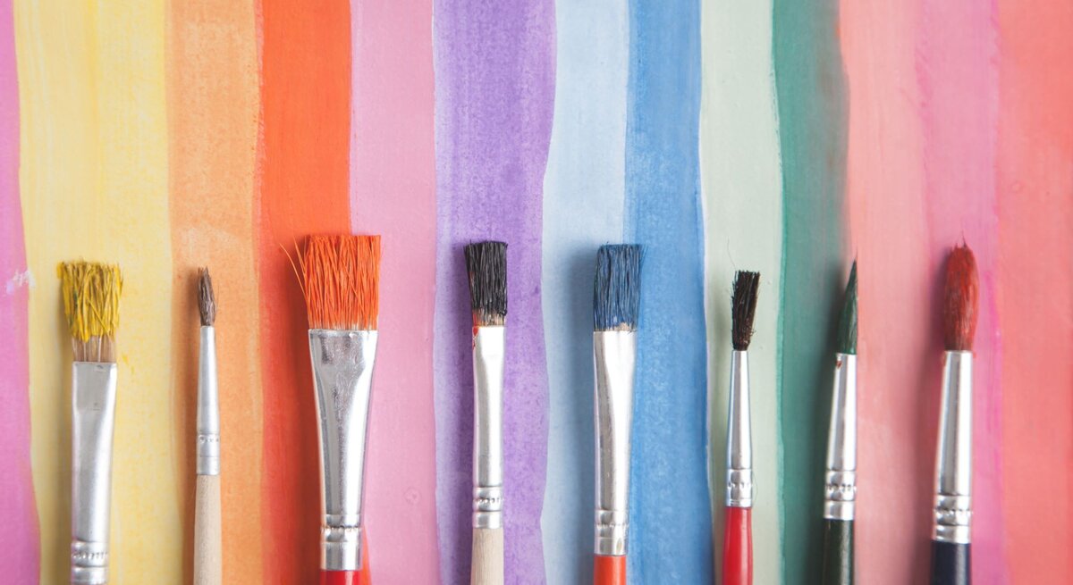 Acht verschiedenfarbige Pinsel liegen nebeneinander vor einem Hintergrund aus bunten Aquarellstreifen in Regenbogenfarben.