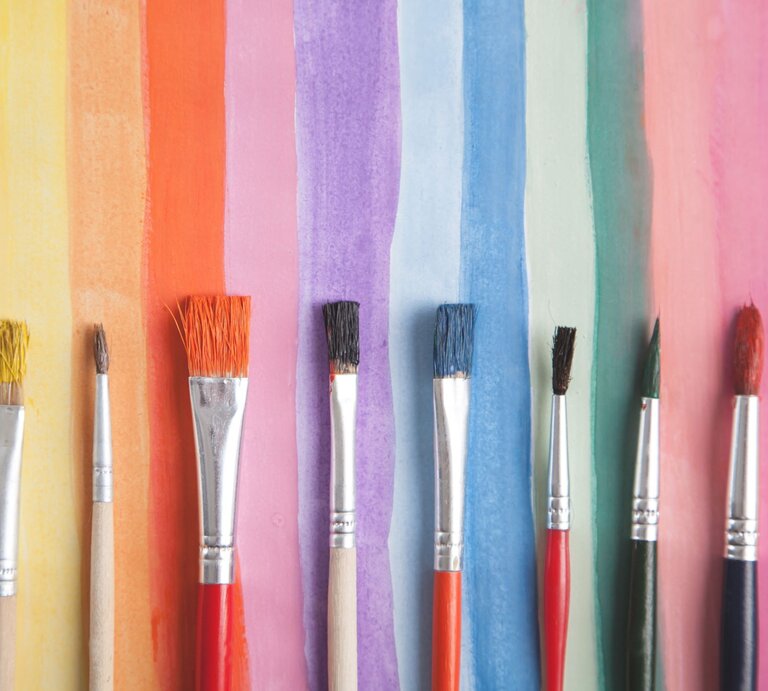 Acht verschiedenfarbige Pinsel liegen nebeneinander vor einem Hintergrund aus bunten Aquarellstreifen in Regenbogenfarben. | © Adobe Stock/andranik123