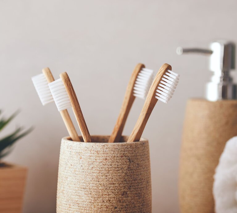 Vier Bambuszahnbürsten mit weißen Borsten stehen in einem beigefarbenen Halter vor einem schlichten Hintergrund mit Seifenspender. | © Adobe Stock/Rostislav Sedlacek