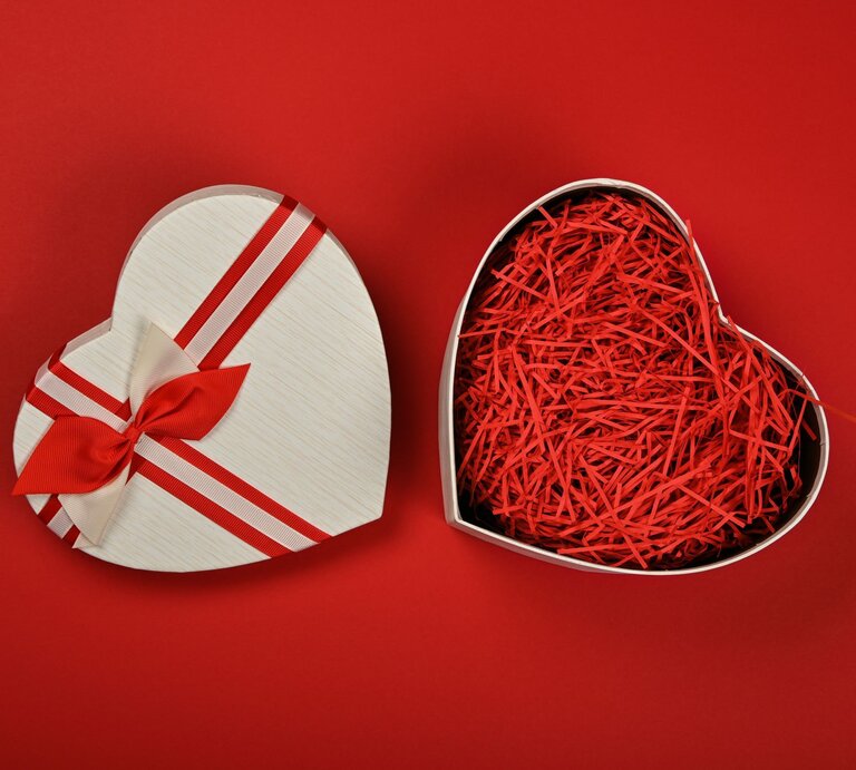 Geöffnete herzförmige Geschenkbox mit rotem Papierstreifen-Füllmaterial auf rotem Hintergrund, der Deckel mit Schleife liegt daneben. | © Adobe Stock/breakingthewalls