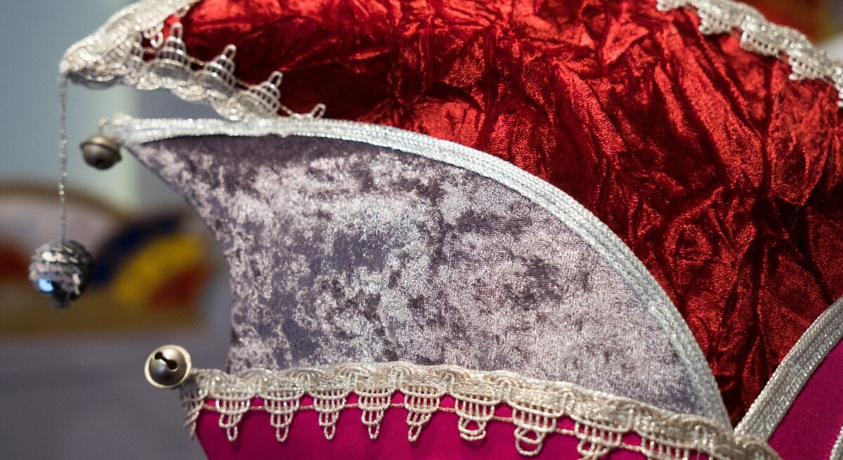 Nahaufnahme einer detailreichen Narrenkappe aus rotem Samt und grauem Stoff, verziert mit goldener Spitze und kleinen Glöckchen.