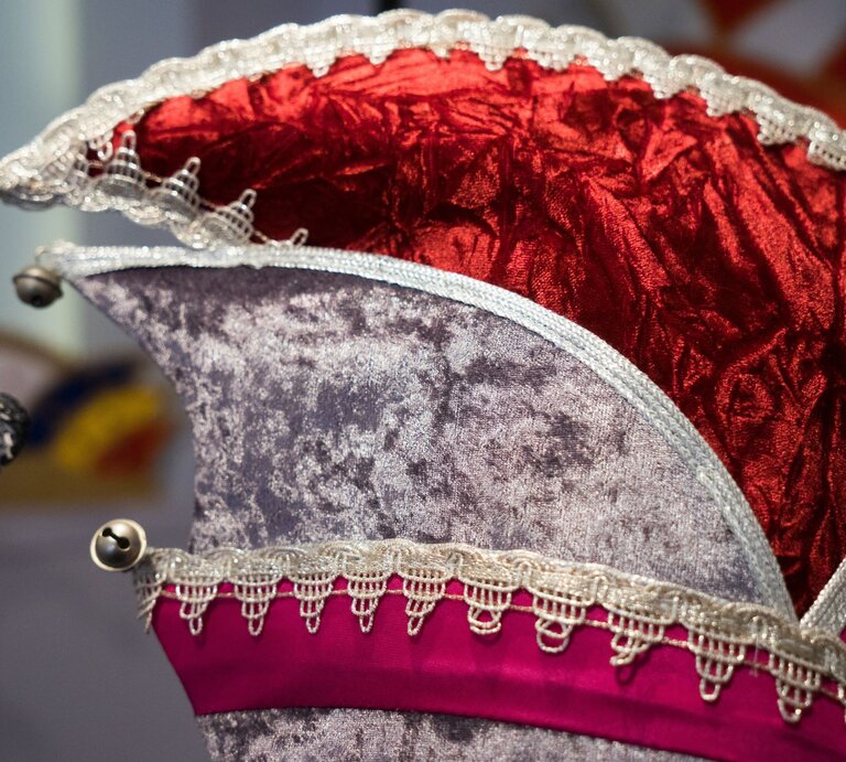 Nahaufnahme einer detailreichen Narrenkappe aus rotem Samt und grauem Stoff, verziert mit goldener Spitze und kleinen Glöckchen. | © Adobe Stock/Cezanne-Fotografie