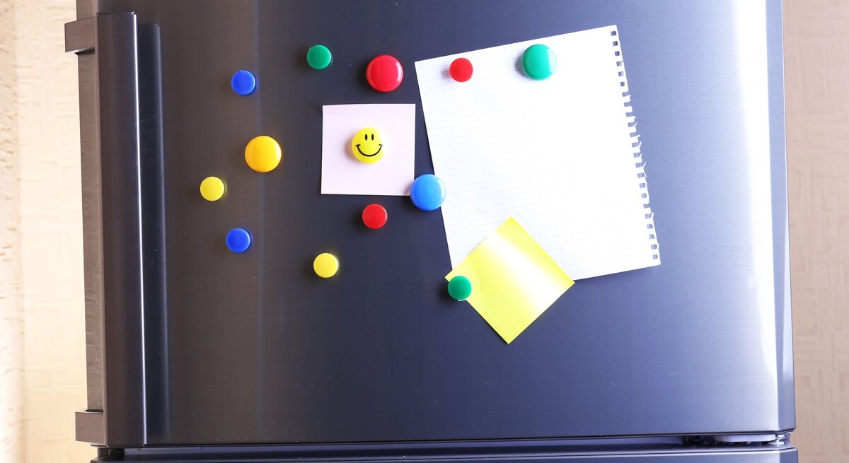 Kühlschranktür aus Edelstahl mit bunten Magneten, darunter ein Smiley-Magnet, und Notizzetteln im warmen Tageslicht.