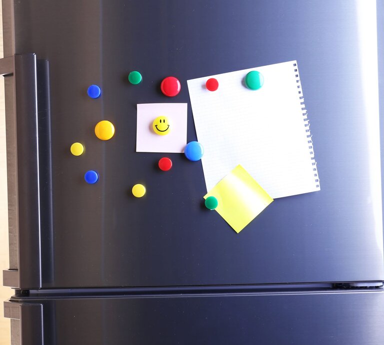 Kühlschranktür aus Edelstahl mit bunten Magneten, darunter ein Smiley-Magnet, und Notizzetteln im warmen Tageslicht. | © Adobe Stock/Africa Studio