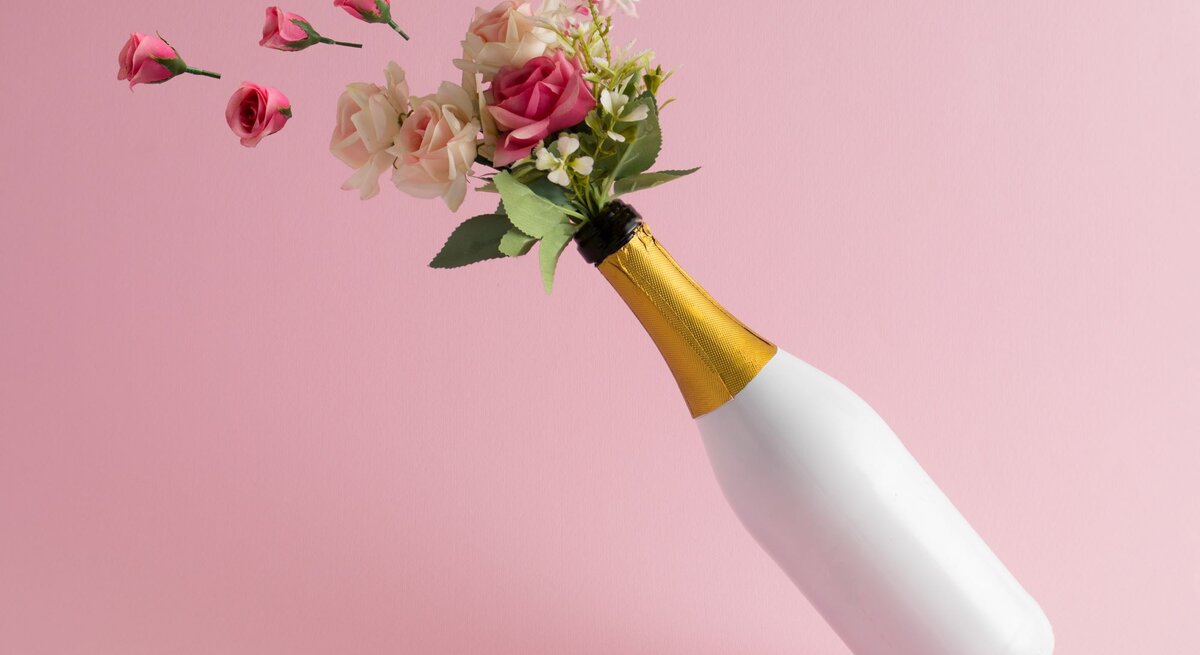 Ein kreatives Blumenbouquet mit rosa und cremefarbenen Rosen in einer Sektflasche auf minimalistischem rosa Hintergrund.