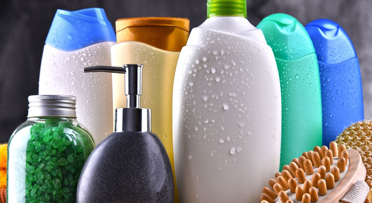 Verschiedene bunte Duschgel- und Shampoo-Flaschen mit Wassertropfen, ein Glas mit grünen Badesalzkristallen und eine Holzmassagebürste auf dunklem Hintergrund.