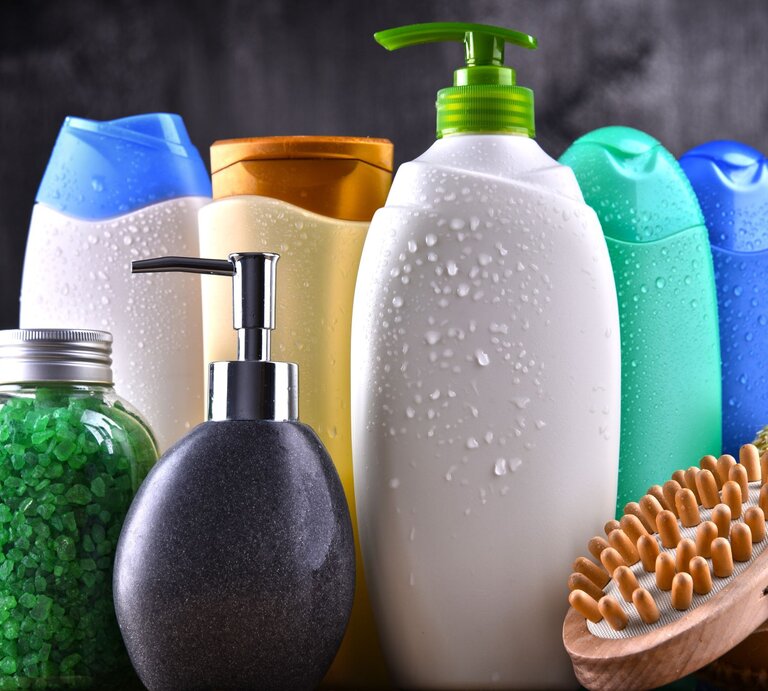 Verschiedene bunte Duschgel- und Shampoo-Flaschen mit Wassertropfen, ein Glas mit grünen Badesalzkristallen und eine Holzmassagebürste auf dunklem Hintergrund. | © Adobe Stock/monticellllo