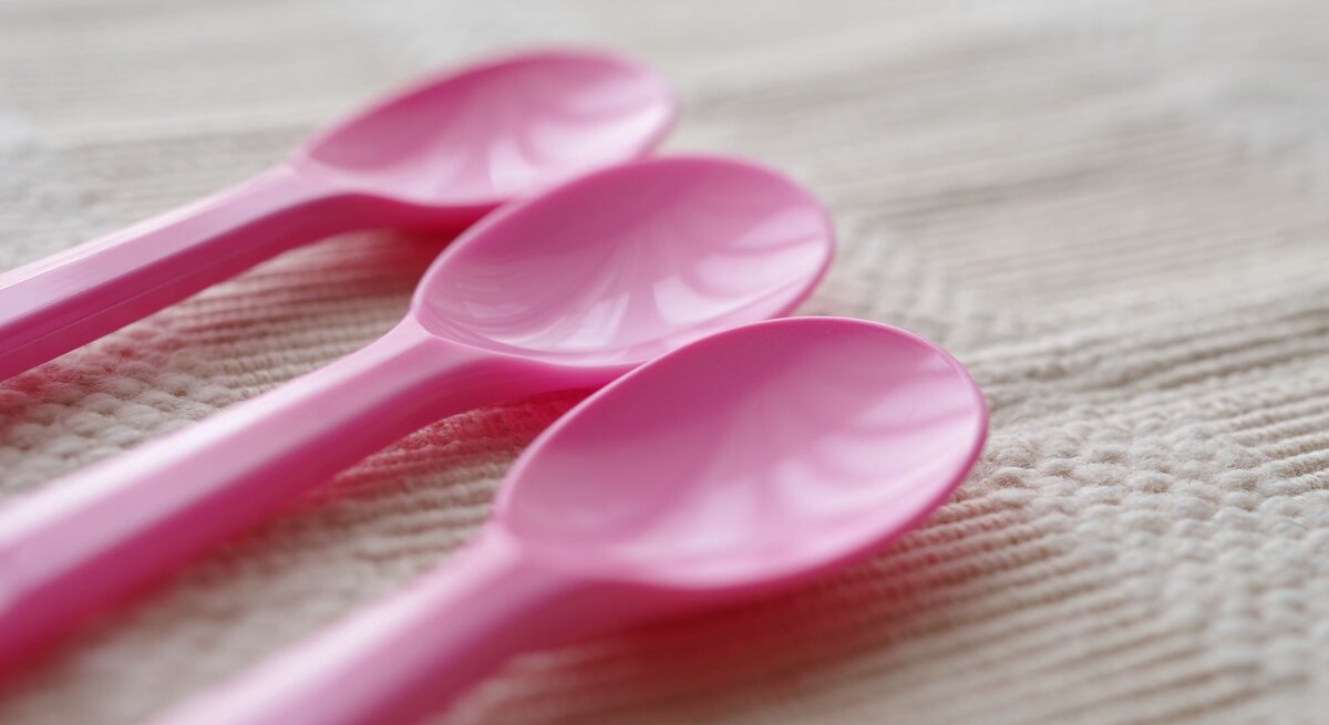 Drei rosa Plastiklöffel liegen nebeneinander auf einer cremefarbenen, strukturierten Stoffoberfläche im weichen Tageslicht.