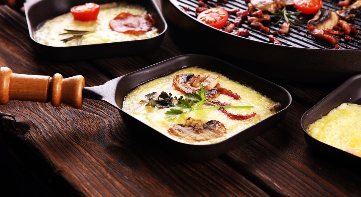 Zwei Raclette-Pfännchen mit geschmolzenem Käse, Speck, Pilzen und Tomaten auf dunklem, selbst gebautem DIY-Raclette-Board. 