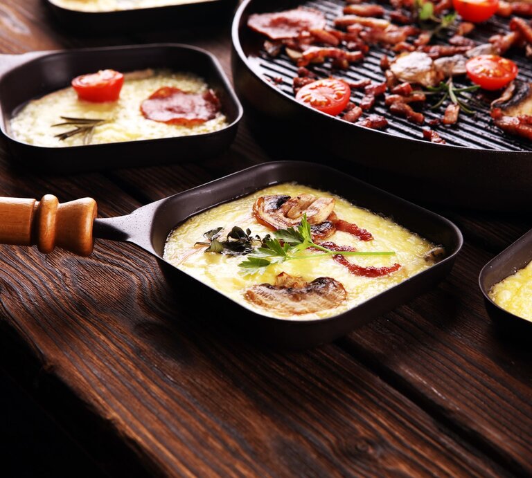 Zwei Raclette-Pfännchen mit geschmolzenem Käse, Speck, Pilzen und Tomaten auf dunklem, selbst gebautem DIY-Raclette-Board.  | © AdobeStock/Roland