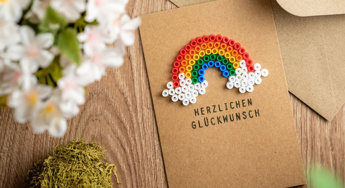 Glückwunschkarte aus braunem Papier mit Regenbogen aus bunten Bügelperlen, umgeben von Blumen und Moos auf Holztisch.