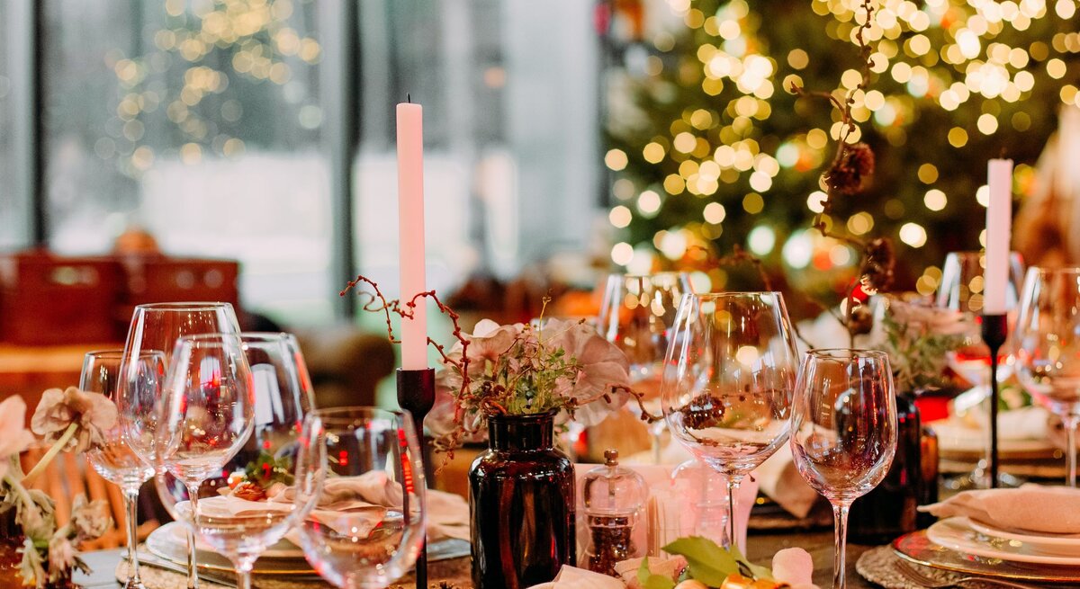 Festlich gedeckter Tisch mit eleganter Weihnachtsdeko aus goldenen Serviettenringen, Kerzen und Tannenzapfen vor leuchtendem Weihnachtsbaum.