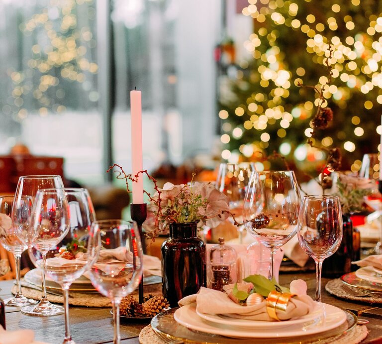 Festlich gedeckter Tisch mit eleganter Weihnachtsdeko aus goldenen Serviettenringen, Kerzen und Tannenzapfen vor leuchtendem Weihnachtsbaum. | © GettyImages/Yuriy Kovtun