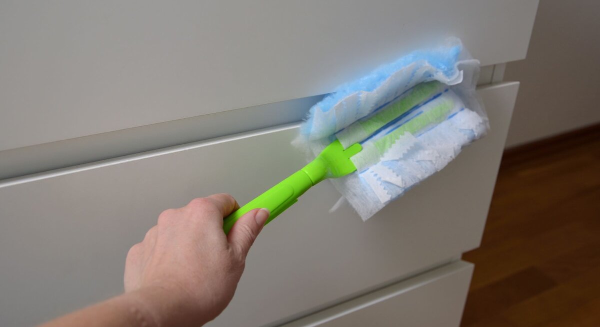 Eine Hand reinigt mit einem grünen Staubwischer und einem blauen Tuch eine weiße Kommode vor Holzfußboden.