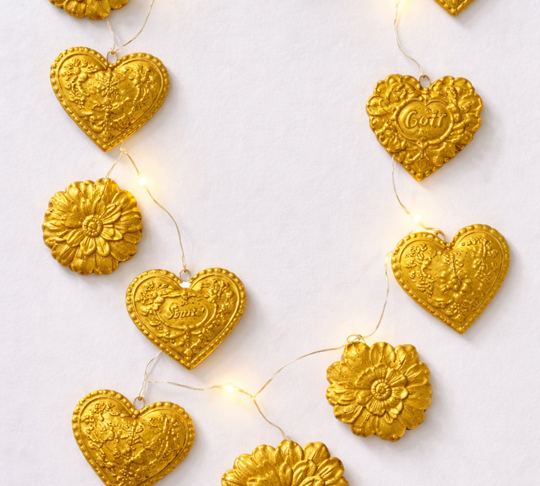 Goldene Herzen und Blumen mit Prägungen hängen an einer Lichterkette auf weißem Hintergrund in festlicher Anordnung. | © Generiert mit KI (Erstellt mit ChatGPT)