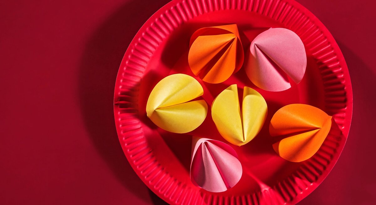 Sechs bunte Papierglückskekse in Gelb, Orange und Rosa liegen auf einem roten Pappteller vor einfarbig rotem Hintergrund.