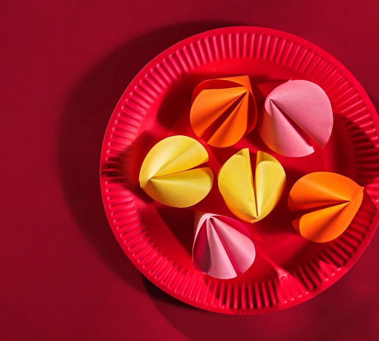 Sechs bunte Papierglückskekse in Gelb, Orange und Rosa liegen auf einem roten Pappteller vor einfarbig rotem Hintergrund. | © Adobe Stock/DyrElena