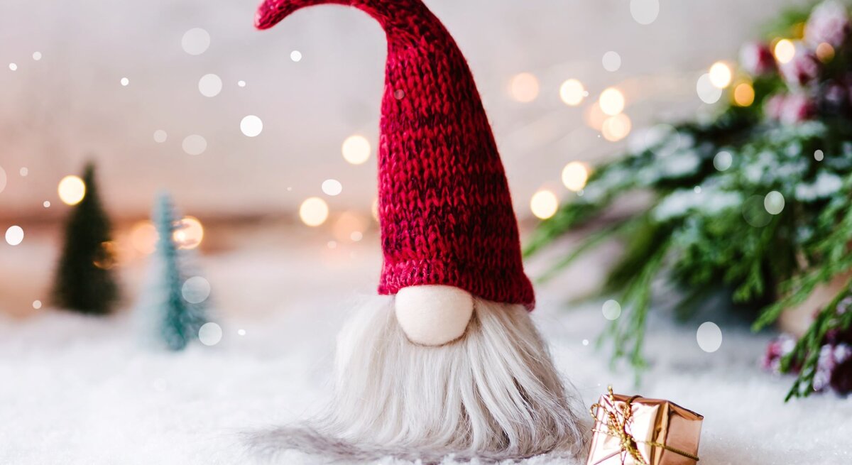 Ein selbst gebastelter Wichtel aus Socken mit roter Strickmütze und grauem Bart steht auf schneebedecktem Boden, daneben ein goldenes Geschenk.